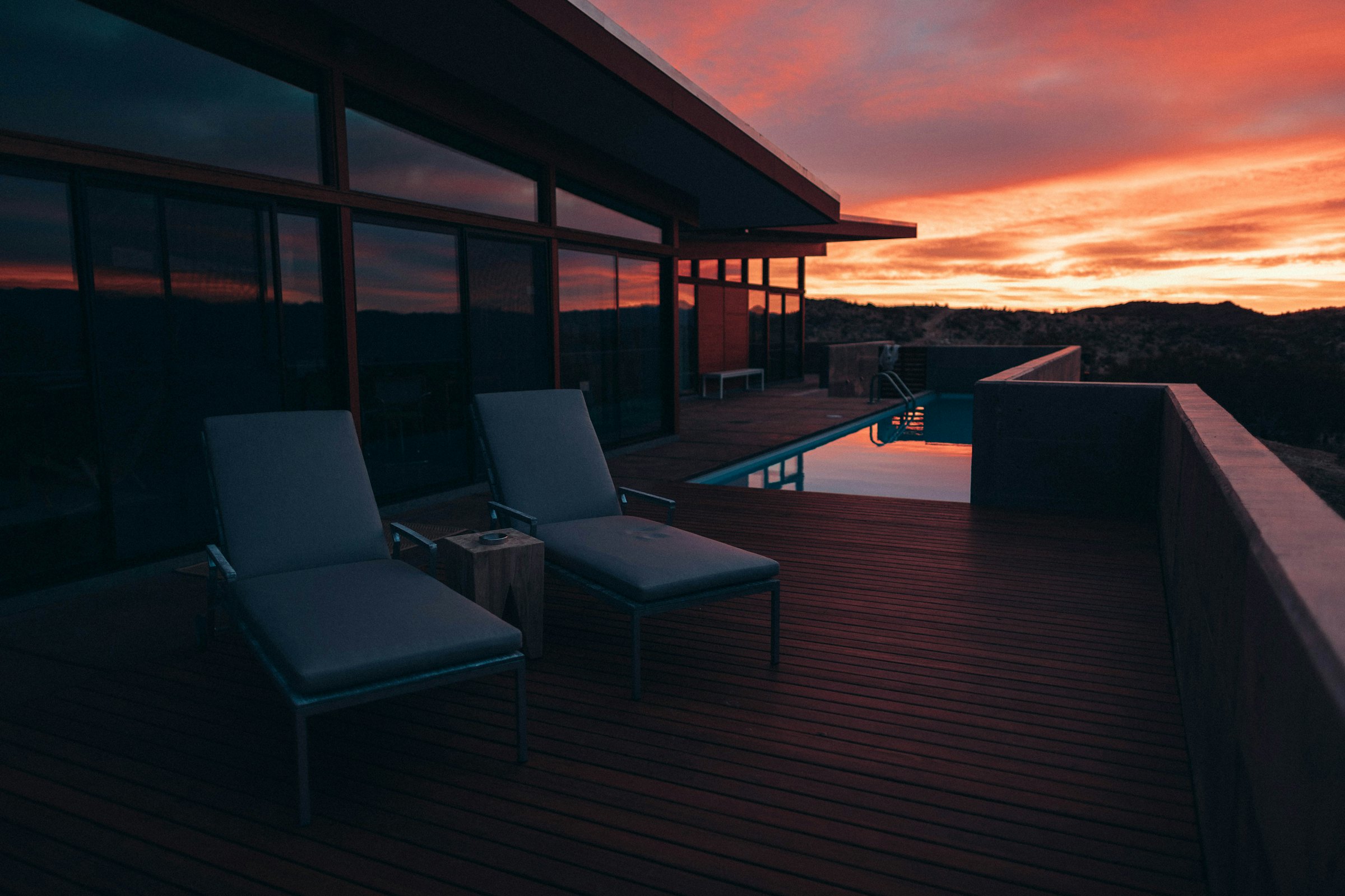 Two lounge chairs on a wooden terrace beside a pool under a red evening sky